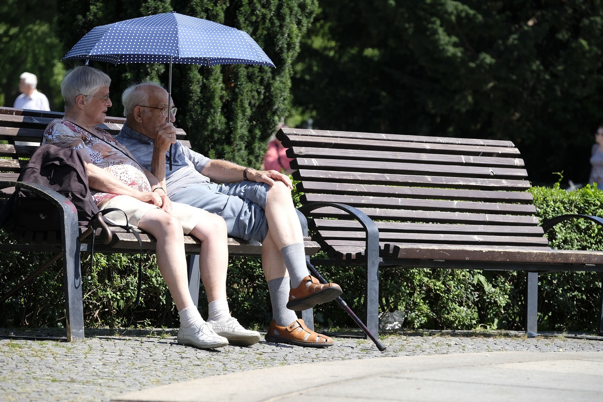 EIn altes Paar sitzt zusammen auf einer Bank mit einem Sonnenschirm in der Hand